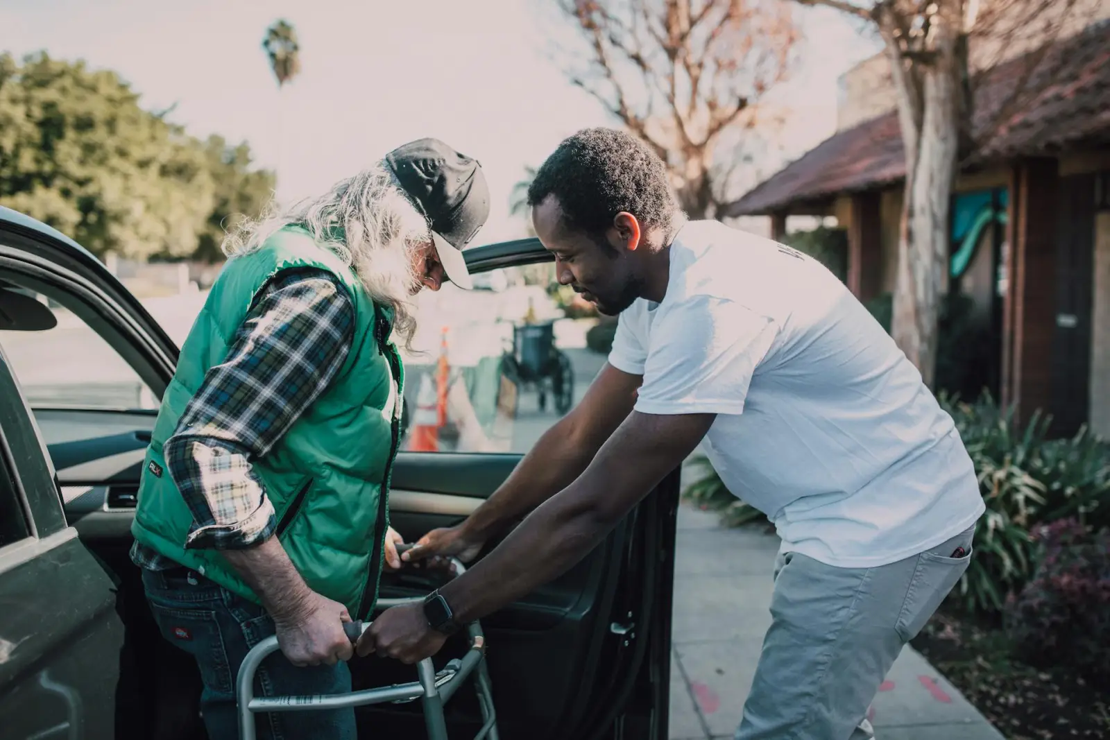 Younger Black man helping an older adult exit a car with a walker in a sunny parking area