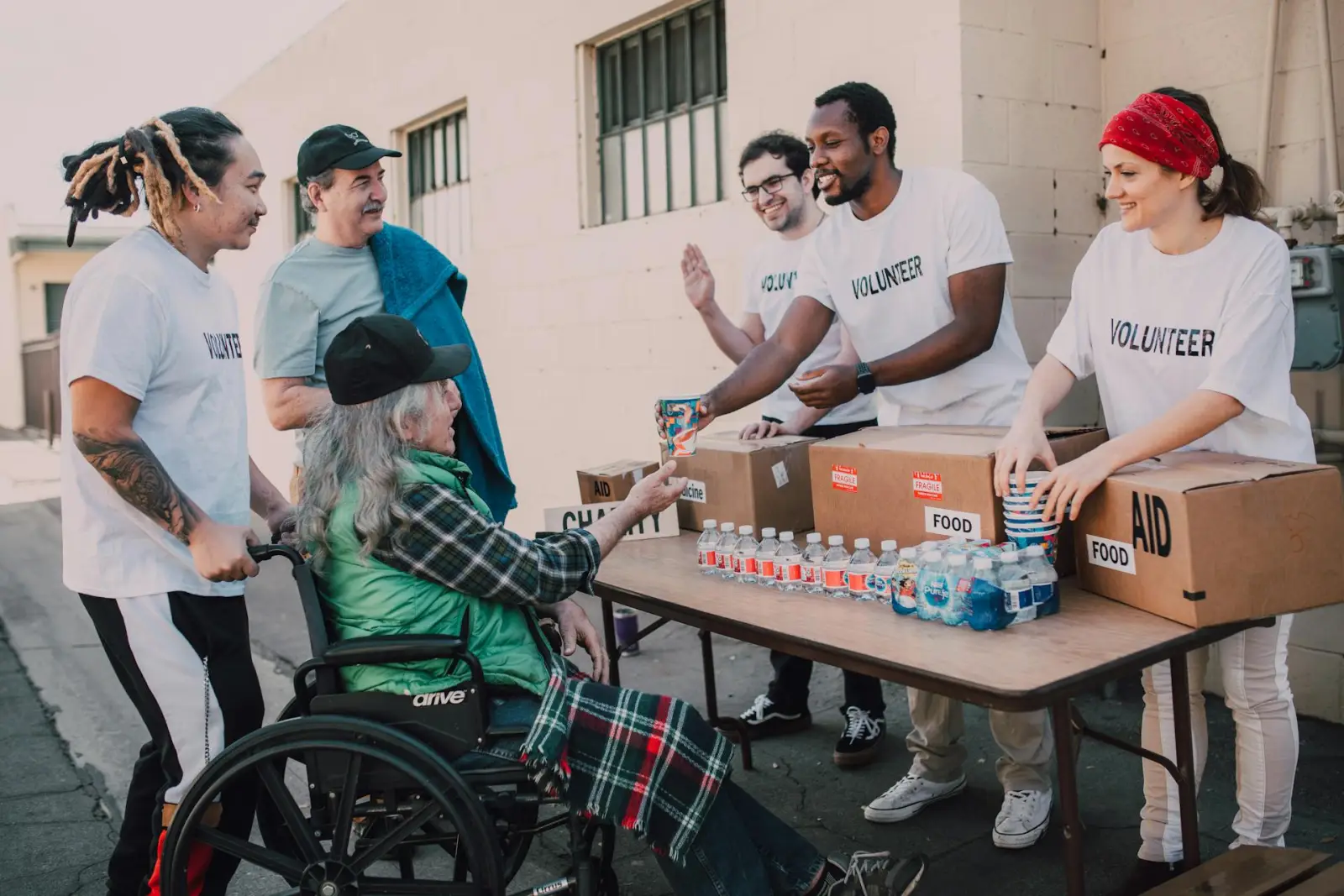 Community volunteers assisting people at an outdoor aid table, including a person using a wheelchair